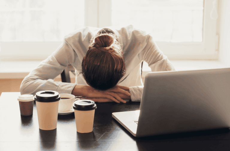 person showing signs of burnout at work with their head resting on a desk and coffee cups nearby.