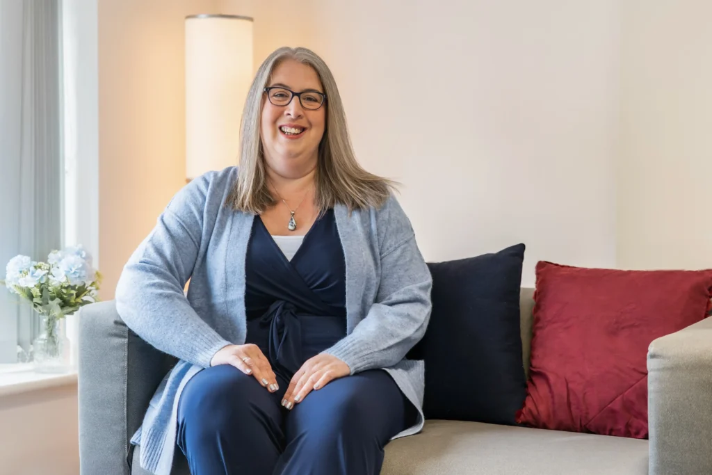 Hannah Paskin sitting comfortably on a sofa, dressed in a blue cardigan and navy outfit.